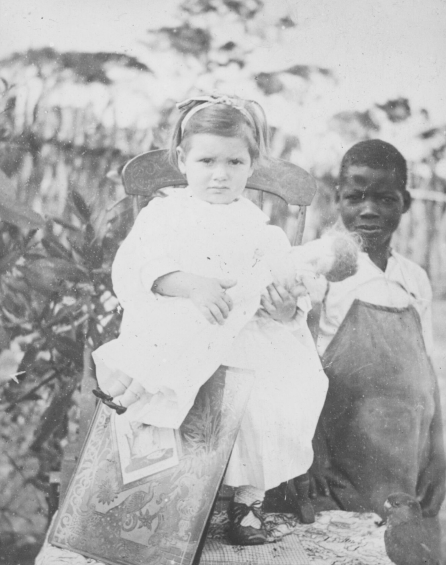 Kind aus Kolonialmacht in weiß gekleidet, erhöht vor einer Einheimischen sitzend.